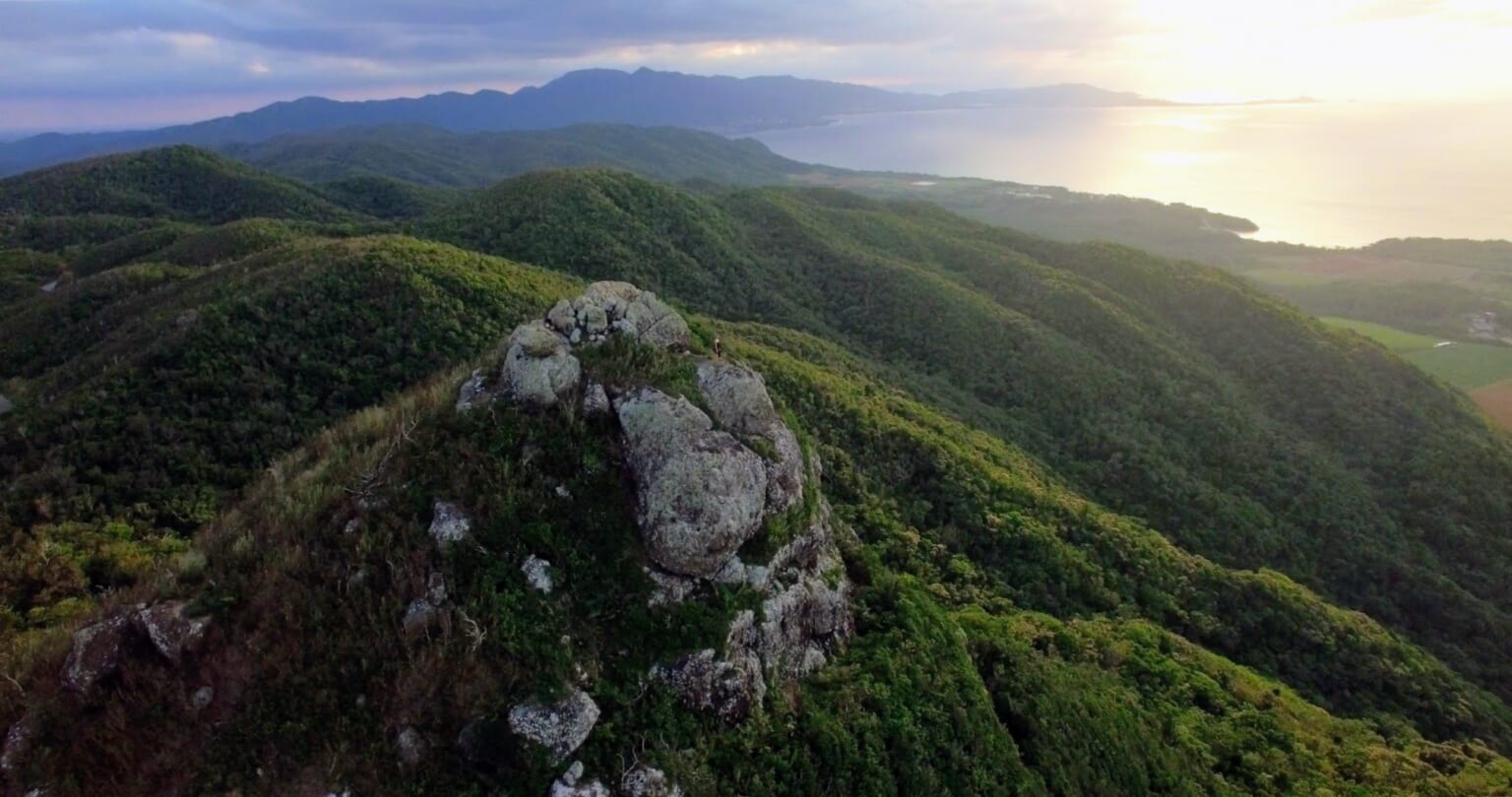The rocky summit of Nosoko Mape above forested hills, with Ishigaki’s coastline and sea beyond.