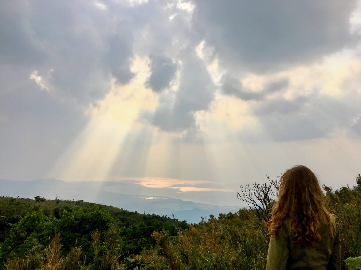 A walker seen from behind on Omotodake, with sunlight breaking through clouds over the sea.